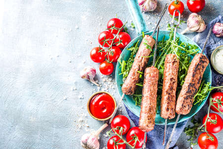 Shish kebab on skewers, from minced beef meat. Lula kebab, traditional Caucasian recipe. On blue concrete background, with sauces,  tomatoes and veggie salad top view copy spaceの写真素材