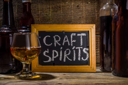 Craft spirit alcohol. Various bottles and glass with hand crafted hard strong alcohol drinks. Old wooden rustic background copy spaceの写真素材
