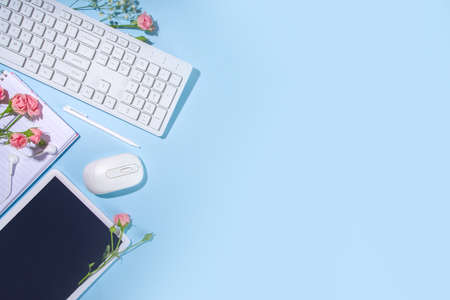 Spring office desk flatlay with keyboard, notepad, tablet, mouse, headphones, spring flowers and leaves, high-colored bright sunny blue background, top view frameの写真素材