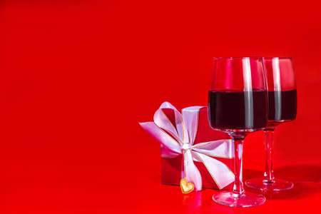 Valentine day gift box and wine glasses, with gold heart decor over red background copy spaceの写真素材