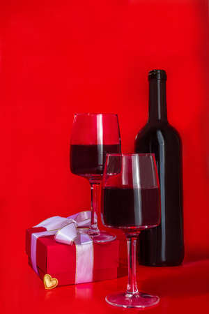 Valentine day gift box and wine glasses, with gold heart decor over red background copy spaceの写真素材