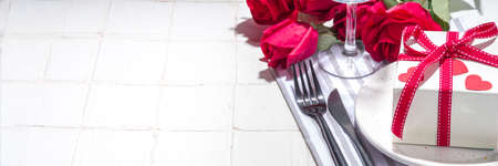 Valentine day table setting with plate, gift box with festive red ribbon, wine glass, fork and knife, red roses flowers bouquet white tiled table flatlay copy spaceの写真素材