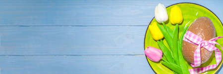 Easter holiday table setting with tulip flowers, big chocolate egg and festive ribbon, top view on...