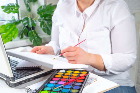 Online learning, hobby classes concept. Girl woman faceless close-up angle learns to paint pictures, draw. Hands with a painting, paints and a brush, a laptop in the frameの写真素材