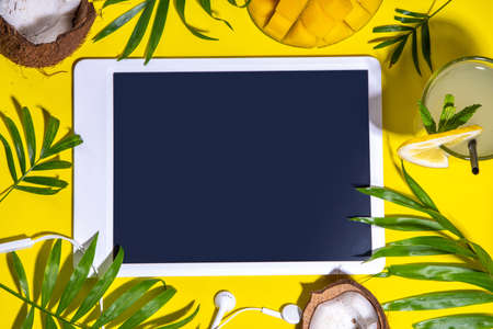 Simple high-colored summer holiday background. Tablet with empty screen with tropical cocktail, summer fruits, palm leaves, on bright yellow background. Travel, vacation flat lay top view copy spaceの写真素材