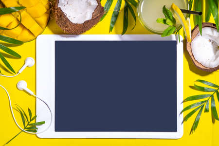 Simple high-colored summer holiday background. Tablet with empty screen with tropical cocktail, summer fruits, palm leaves, on bright yellow background. Travel, vacation flat lay top view copy spaceの写真素材