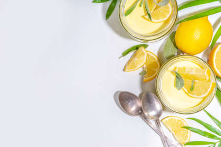 Lemon panna cotta dessert with fresh lemon wedges and sage leaves. Yellow citrus fruit panna cotta in portion jars on a white backgroundの写真素材