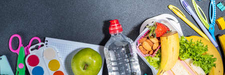 Healthy school lunch box: sandwich, vegetables, fruit, nuts and yogurt with school kids supplies, accessories and backpack on black background flatlay copy space. Back to school conceptの写真素材