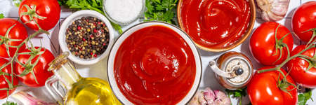 Homemade ketchup sauce. Two bowl with tomato ketchup red sauce with ingredient for cooking - herbs, raw fresh tomatoes, olive oil, garlic, salt, spices, on white tiled background top view copy spaceの写真素材