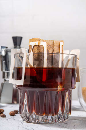 Trendy instant brewed drip coffee, cold brew coffee. Drip filter coffee bag in glass cup on white table, with brown sugar, coffee beans, pot with hot waterの写真素材