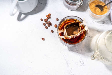 Trendy instant brewed drip coffee, cold brew coffee. Drip filter coffee bag in glass cup on white table, with brown sugar, coffee beans, pot with hot waterの写真素材