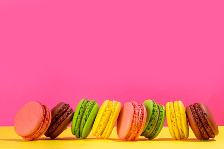 Bright colorful (yellow, pink, green, brown) various flavor macarons sweet cookies on high-colored pink yellow background. Stack of small french macaron cakes, copy space flatlayの写真素材