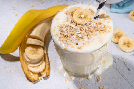 Vegan banana drink, healthy diet snack. Banana oatmeal smoothie in glass, with oats flakes and banana slices, white background copy spaceの写真素材