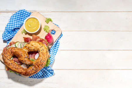 Oktoberfest pretzel bagel sandwich. Oktoberfest day breakfast, burger recipe for holiday beer party. Big pretzel with ham, cheese, fresh vegetables, with radish, beer, checkered tablecloth copy spaceの写真素材