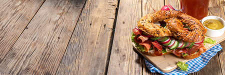 Oktoberfest pretzel bagel sandwich. Oktoberfest day breakfast, burger recipe for holiday beer party. Big pretzel with ham, cheese, fresh vegetables, with radish, beer, checkered tablecloth copy spaceの写真素材