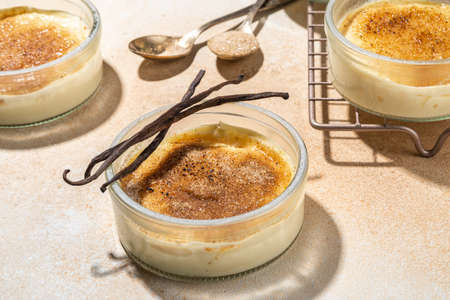 Sweet creme brulee dessert. Portioned glass jars with homemade creme brulee with caramelized sugar crust topping, on beige colored background with sugar in spoon and vanilla copy spaceの写真素材