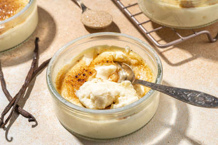 Sweet creme brulee dessert. Portioned glass jars with homemade creme brulee with caramelized sugar crust topping, on beige colored background with sugar in spoon and vanilla copy spaceの写真素材