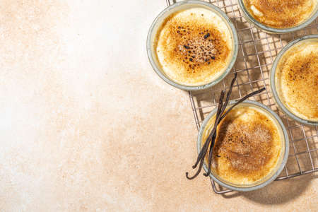 Sweet creme brulee dessert. Portioned glass jars with homemade creme brulee with caramelized sugar crust topping, on beige colored background with sugar in spoon and vanilla copy spaceの写真素材