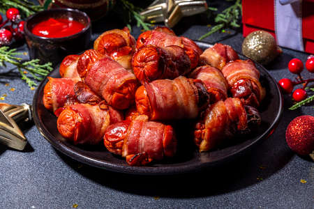 Traditional Christmas dish recipe Pigs in blankets, sausages wrapped in bacon, oven baked puff pastry hot dogs rolls with xmas tree branches, decoration and gifts on black tableの写真素材