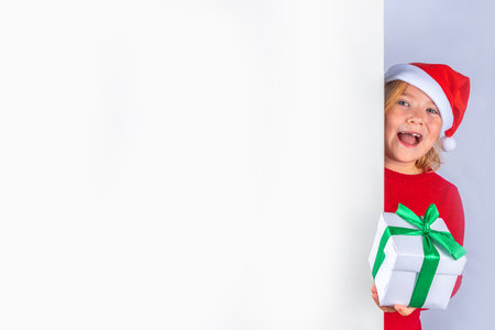 Christmas holiday background with funny blond girl. Little kid girl in red Christmas sweater and funny santa hat costume, with gift box on white background, Xmas greeting card, copy spaceの写真素材