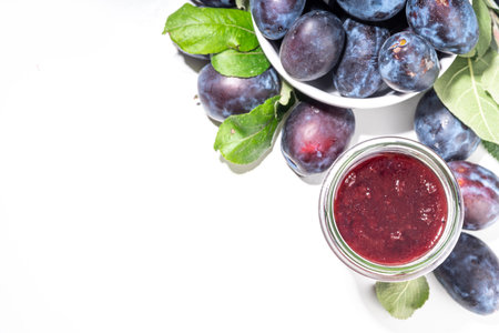 Red blue plum jam in small jar. Homemade autumn plum jam with fresh fruits. Fall preparations and canning on white table background copy spaceの写真素材