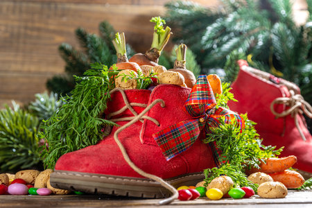 Sinterklaas, Saint Nicholas day background. Little kid red shoe with carrots and sweets, trooigoed, pepernoten, candy. Dutch Christmas holiday on cozy wooden background.の写真素材