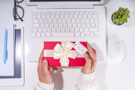 Present from secret santa on workplace table. Office Christmas and New Year celebration game. Office table surface, notebook with festive gift box, white background flatlay copy spaceの写真素材