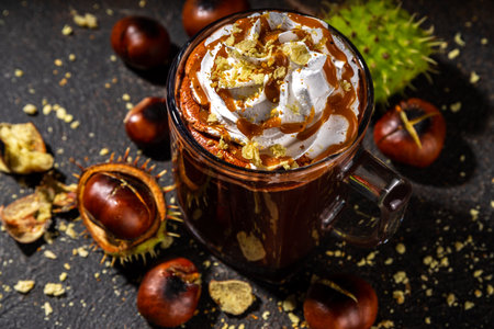 Glass cup with dark chestnut hot chocolate or choco latte, with whipped cream, caramel sauce and roasted chestnuts, over dark table copy spaceの写真素材
