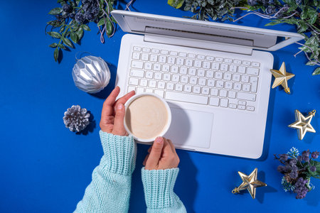 Christmas sale, preparation for holidays concept. winter office workplace background. Laptop, hot chocolate latte cup with Christmas toys, winter tree branches, flat lay on bright blue backgroundの写真素材