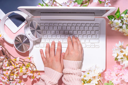 Spring office workplace, blogging flat lay background. White laptop, with headphones, tablet, spring flowers bouquet on pink pearl background, girl's hands typing on a laptop.の写真素材