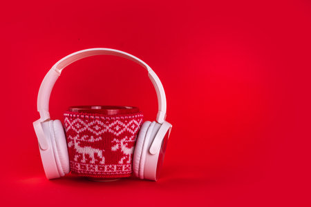 Christmas mood and vibe, warming traditional new year songs music listen concept. Red mug with christmas knitted sweater with big headphones on red background, minimal simple design copy spaceの写真素材