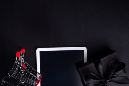 Black friday sale background. Simple black flat lay with sign "Black friday", gift boxes, shopping cart, laptop, tablet, headphones top view copy space. Online Black friday shopping conceptの写真素材