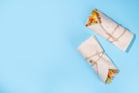 Classic street food shawarma or burrito, healthy sandwich wrapped in tortilla flat bread with fried chicken meat, fresh vegetables, sauce, on bright blue background, top view flat lay copy spaceの写真素材