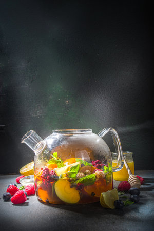 Fruit and berry tea in teapot. Hot drink with lemon, mint, blueberry, ginger, orange, apple. Hot flavored steam beverage on dark blue background copy spaceの写真素材