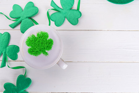 Simple background for St. Patrick's Day. Cup of cappuccino latte coffee with whipped milk foam and decor of green sugar powder shamrock four-leaf clover on white wooden background copy spaceの写真素材