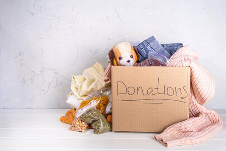 Donation box with clothes, toys and food on white table background. Cardboard donation box full with...