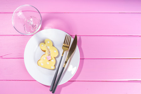 Cute Easter table setting, spring holiday decorated table with plate, glass, cutlery, tulip flowers bouquet, Easter decoration symbols and treats, on pink wood bright table, top view copy spaceの写真素材
