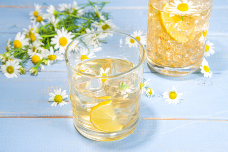 Herbal organic cold drink. Infused flower water. Iced daisy Chamomile tea or lemonade with lemon slice and ice cubes, in two different glasses, blue wooden background copy spaceの写真素材