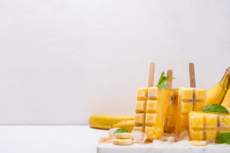Frozen banana fruit lollypops, with ice and mint decor, white kitchen background. Summer cold vegetarian healthy dessert, copy spaceの写真素材