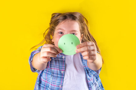 Mental health day, Protecting children from stress concept. Positive thinking, childhood depression therapy. Cute european pre-teenage girl holds paper smiley emoticon in her hand on yellow backgroundの写真素材