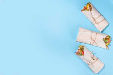 Classic street food shawarma or burrito, healthy sandwich wrapped in tortilla flat bread with fried chicken meat, fresh vegetables, sauce, on bright blue background, top view flat lay copy spaceの写真素材