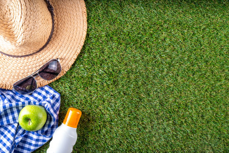 Spring, summer picnic, outdoor recreation background. A woman's straw hat, a picnic blanket or tablecloth, sunscreen spray, an apple on a background of green artificial grass or lawn, copy spaceの写真素材