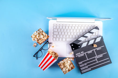 Cinema, film and movie night watching concept. Clapperboard, striped popcorn bucket boxes, with glasses and white laptop on light blue background, copy spaceの写真素材