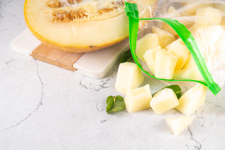 Frozen melon slices, cubes in freezing bags with fresh ripe raw melon on kitchen white table, Healthy simple canning, freezing stock up of fruit harvest for winter concept.の写真素材