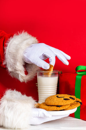 Hand of Santa Claus with traditional Christmas cookie snack with milk glass. Santa Dunking Cookie, on bright festive red and white wooden  background. Merry Christmas and Happy New Year greeting cardの写真素材