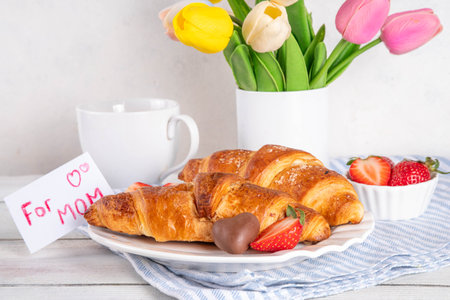 Motherâs Day breakfast background featuring croissant, strawberries, chocolates, coffee mug, bouquet of tulip flowers, as surprise and gift, with greeting card "for mom".の写真素材