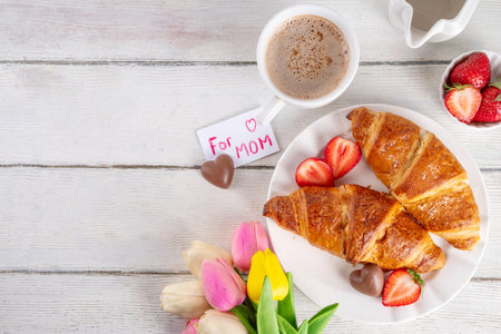 Motherâs Day breakfast background featuring croissant, strawberries, chocolates, coffee mug, bouquet of tulip flowers, as surprise and gift, with greeting card "for mom".の写真素材