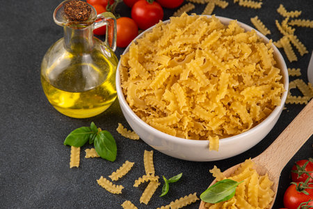 Raw uncooked Mafaldine pasta with ingredients for cooking. Short italian Mafaldine pasta with olive oil, cherry tomatoes, basil, spices on black kitchen table, background copy spaceの写真素材