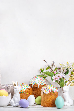 Tasty Glazed Easter cakes with sugar decor, colorful eggs basket, ceramic rabbits and spring flowers on white kitchen table, copy space. Happy Easter Holidays greeting card background copy spaceの写真素材