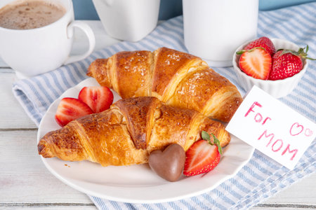 Motherâs Day breakfast background featuring croissant, strawberries, chocolates, coffee mug, bouquet of tulip flowers, as surprise and gift, with greeting card "for mom".の写真素材
